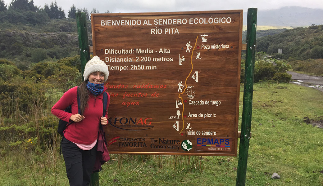 Lady in red jacket and white woolly hat holding on to her backpack straps in front of a trail map to the Pita Canyon, Cotopaxi, Ecuador 