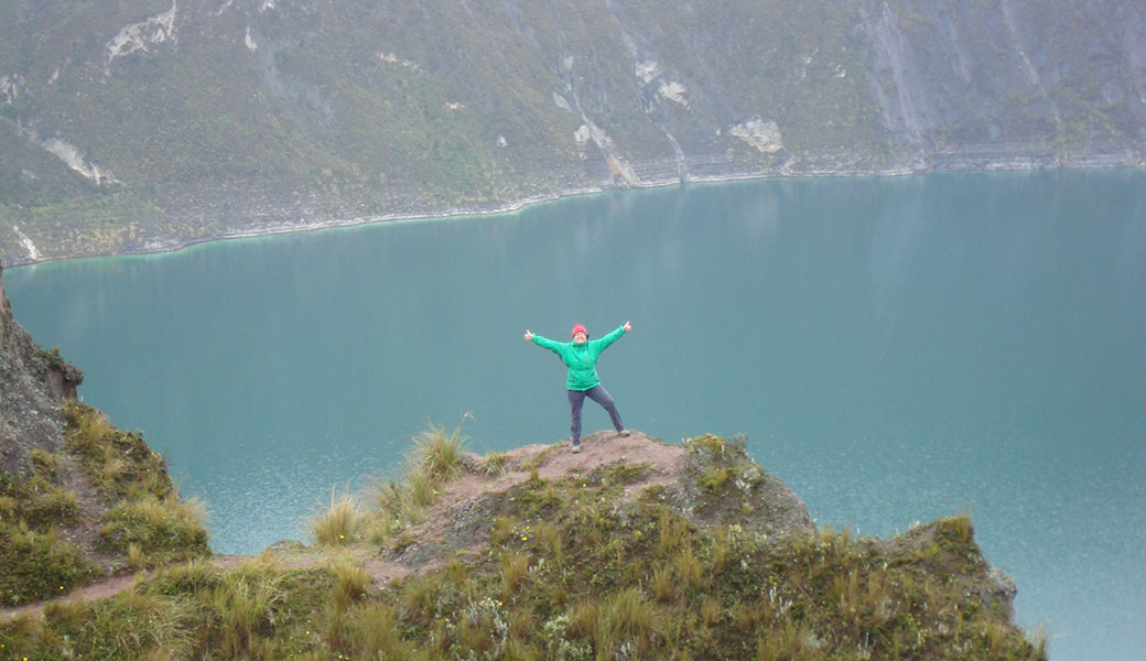 Lady in green jacket with arms in air standing on a rock in front of Quilotoa Crater Lake, Ecuador