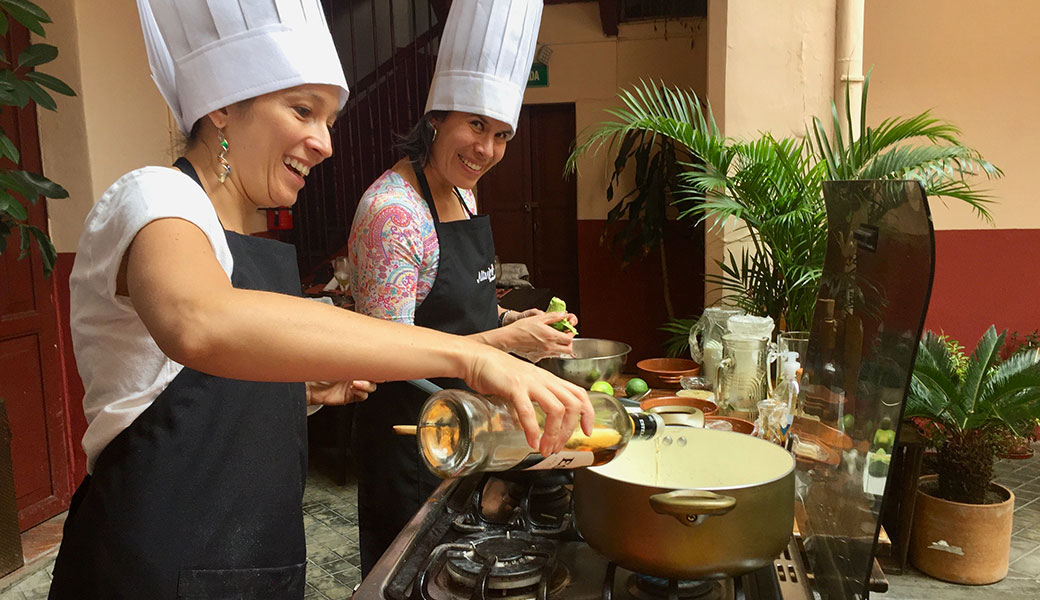 Smiling lady in chef's hat and apron pours rum into boiling saucepan as her smiling friend also in chef's hat and apron looks on